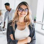 Usměvavá žena s dlouhými blond vlasy a brýlemi stojí se zkříženýma rukama ve světlé kanceláři, zatímco v pozadí pracují její kolegové.