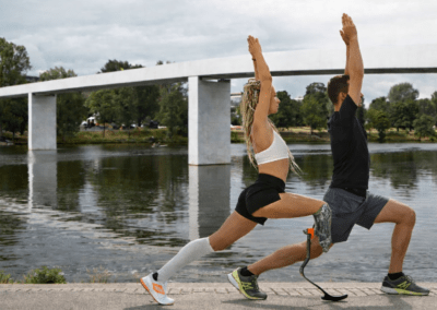 Dva lidé cvičí venku u řeky, oba v pozici výpadu se zdviženýma rukama. Muž má protézu nohy. V pozadí je pod zataženou oblohou vidět moderní bílý most a zeleň.