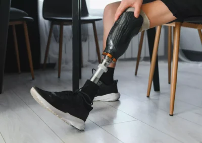 Osoba v černých sportovních botách sedí na židli a ukazuje protézu nohy z uhlíkových vláken. Podlaha je ze světlého dřeva a v pozadí je několik prázdných židlí.