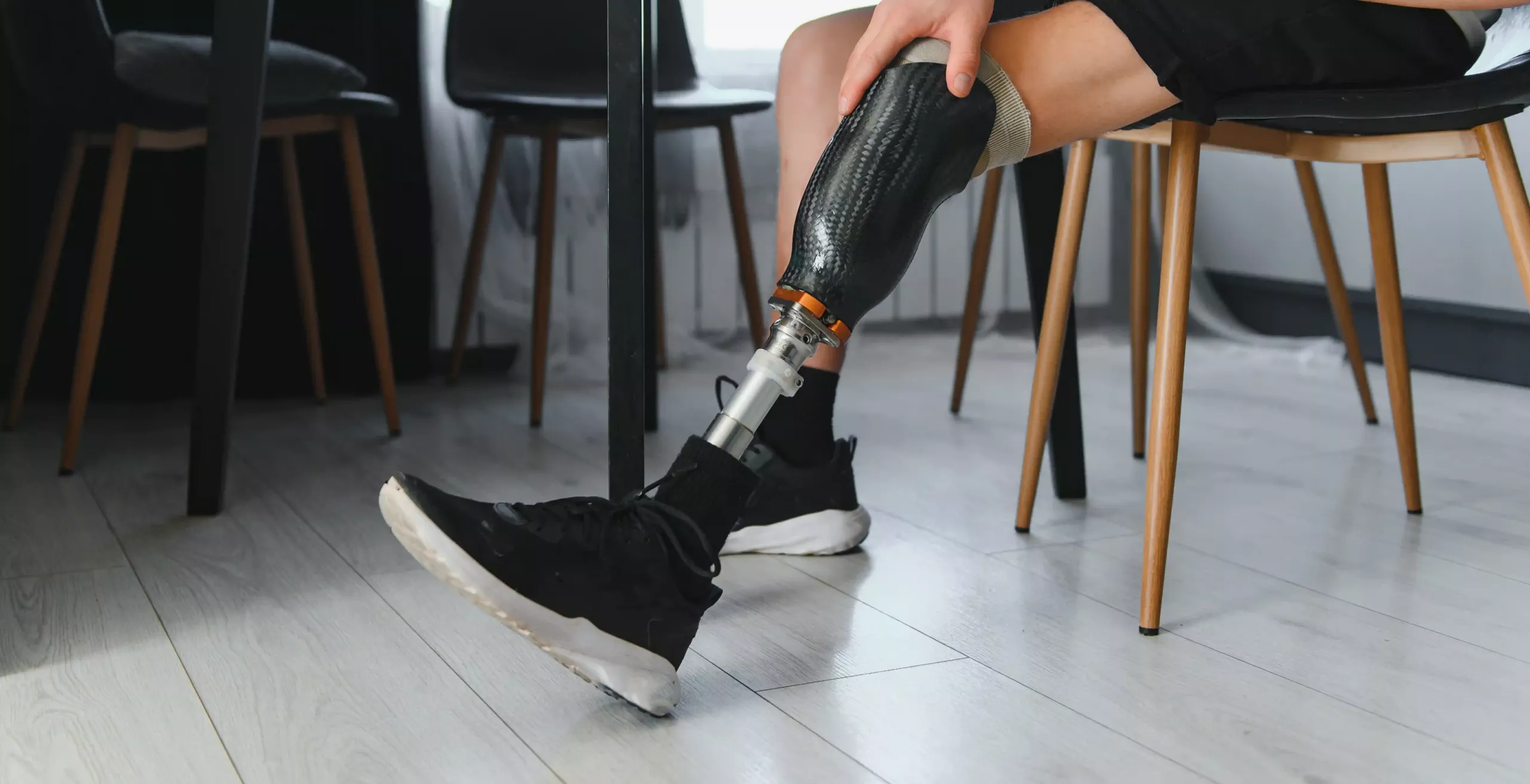 Osoba v černých sportovních botách sedí na židli a ukazuje protézu nohy z uhlíkových vláken. Podlaha je ze světlého dřeva a v pozadí je několik prázdných židlí.