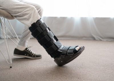 Osoba v šedých teplácích sedí na židli s jednou nohou nataženou a na zraněné noze má černou zdravotní botu; na druhé noze má černou tenisku.