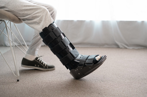 Osoba v šedých teplácích sedí na židli s jednou nohou nataženou a na zraněné noze má černou zdravotní botu; na druhé noze má černou tenisku.