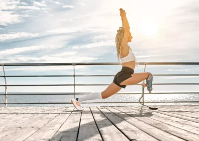 Žena s protézou nohy cvičí na promenádě u oceánu výpadový strečink, má na sobě aktivní oblečení a zvedá ruce nad hlavu ve slunečním světle.